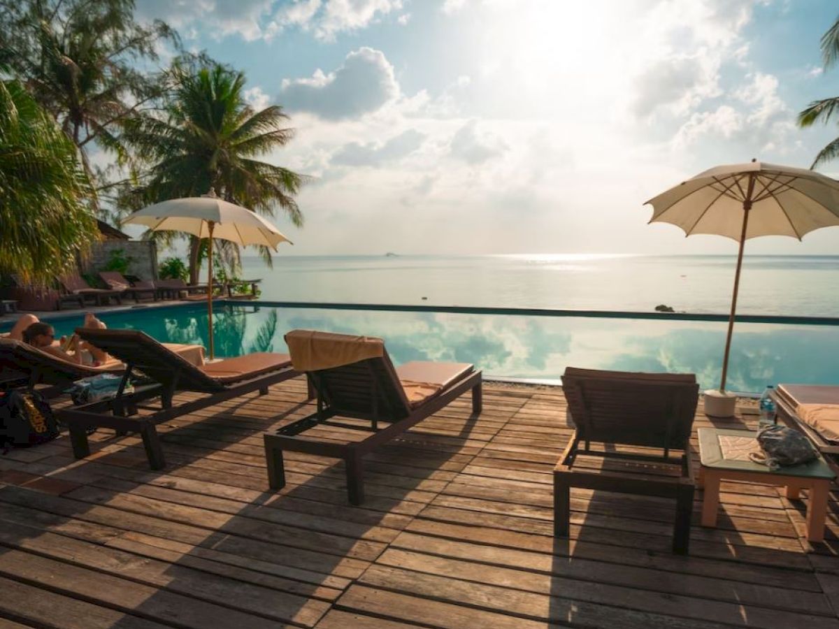 Lounge chairs and umbrellas by a poolside overlook a scenic view of the ocean beneath a partly cloudy sky.