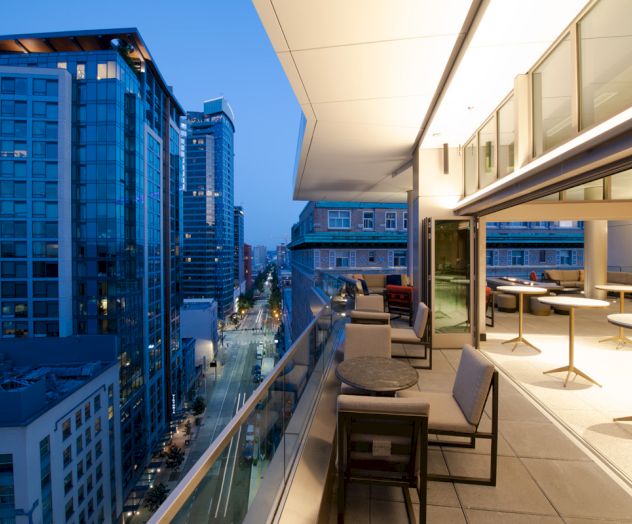 A modern rooftop terrace overlooking a cityscape with tall buildings, featuring seating areas and ambient lighting during evening twilight.