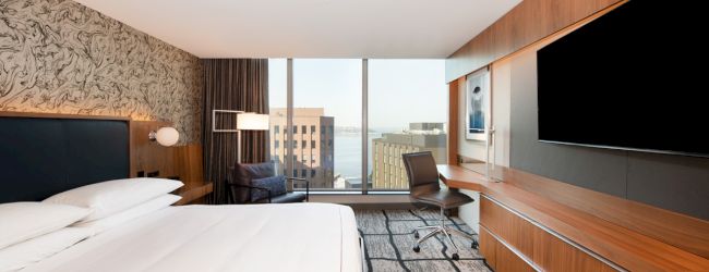 A modern hotel room features a large bed, wall-mounted TV, desk, and floor-to-ceiling windows with a city view.