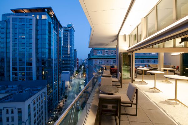 A rooftop patio with modern furniture overlooks city buildings at dusk, featuring a mix of glass and concrete architecture along a quiet street.
