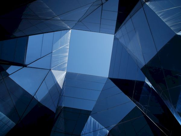 The image shows an upward view of a geometric structure made of reflective glass panes, creating a pattern around a central blue sky.