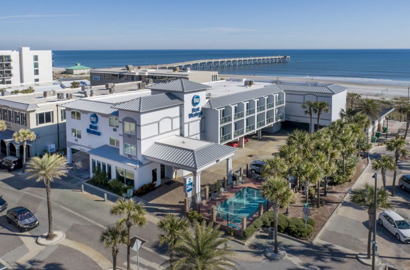 Aerial view of a coastal town with a pier, buildings, palm trees, and parked cars near the beach under a clear blue sky.