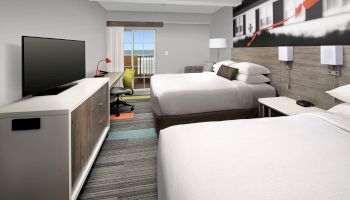 A modern hotel room with two beds, a TV, desk, and balcony view, featuring stylish decor and lighting.