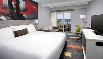 A modern hotel room features a large bed, desk, chair, sofa, and wall art, with a window view and contemporary decor.