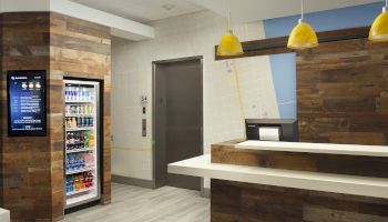 The image shows a modern lobby with a wooden reception desk, a beverage fridge, elevator doors, and pendant lights above the counter.