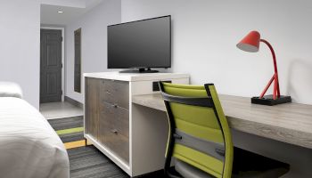 A modern hotel room includes a bed, TV on a cabinet, desk with a red lamp, and a green chair on a colorful carpet.