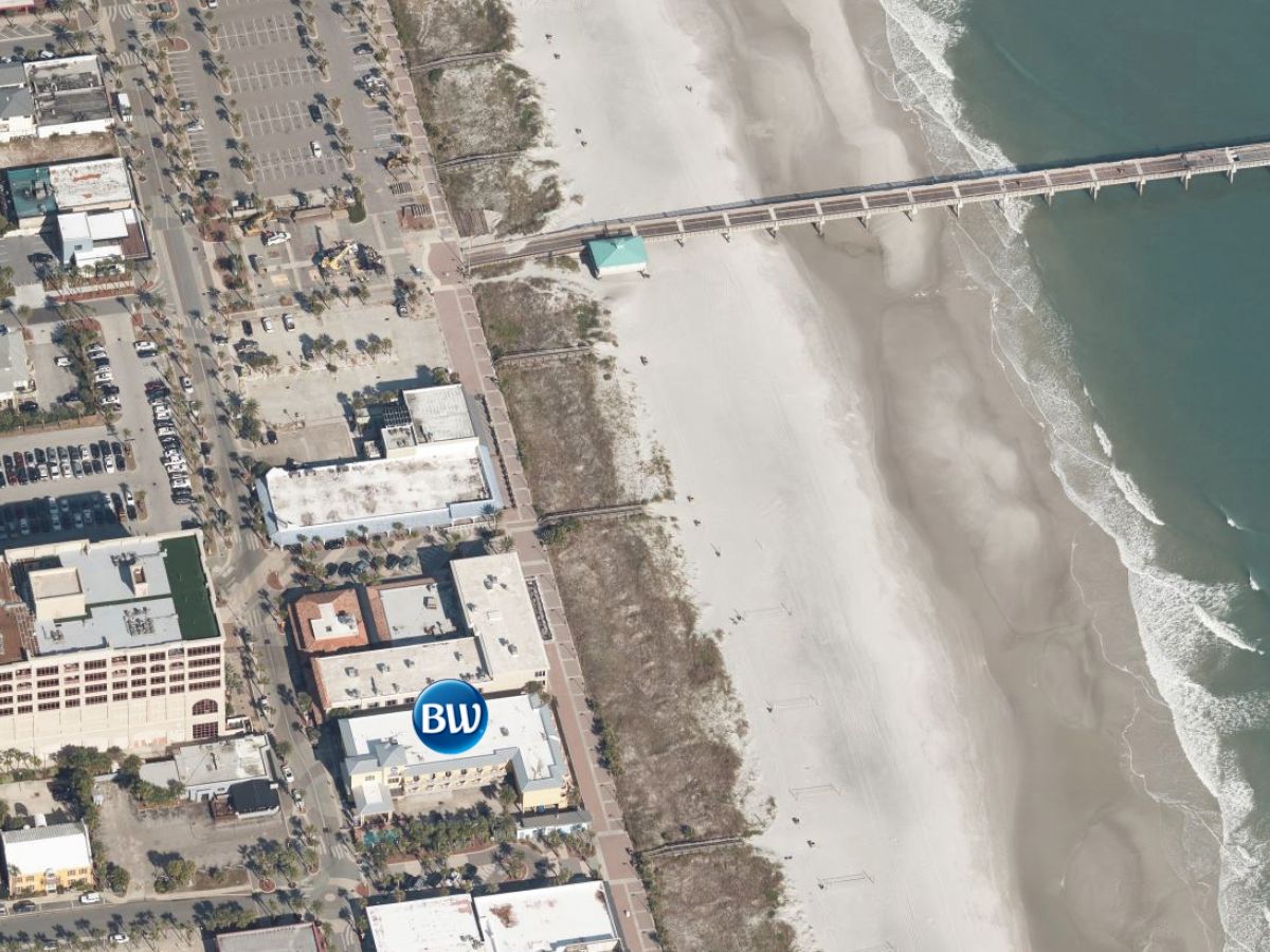 An aerial view of a coastal area shows a pier extending into the ocean, a sandy beach, and buildings near the shoreline.