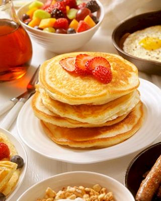 A breakfast spread with pancakes topped with strawberries, syrup, waffles, sausages, eggs, oatmeal, and a bowl of fruit.