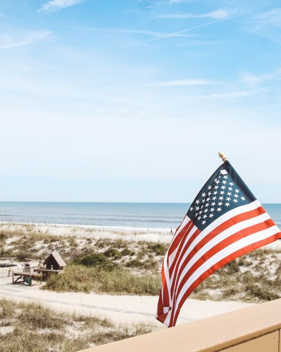 An American flag waves near a sandy beach with grass-covered dunes under a blue sky with scattered clouds in the background.