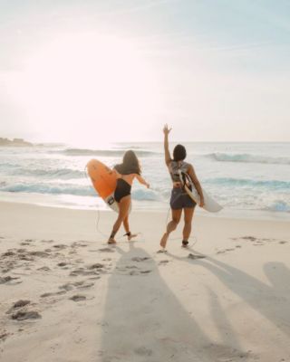Two people walk towards the ocean with surfboards on a sunny beach, casting long shadows on the sand.