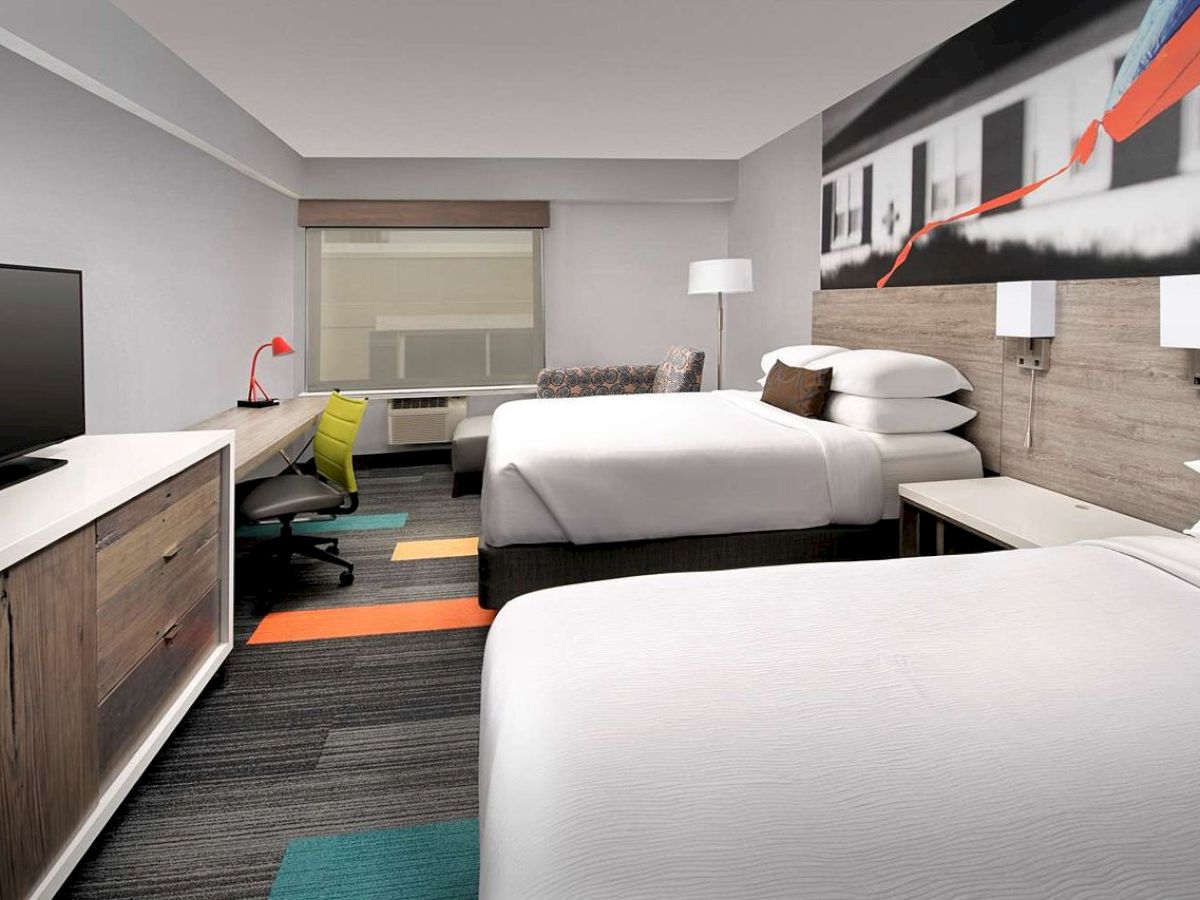 A modern hotel room with two beds, a TV, a desk with a chair, and colorful accents. A large wall art decorates the room.