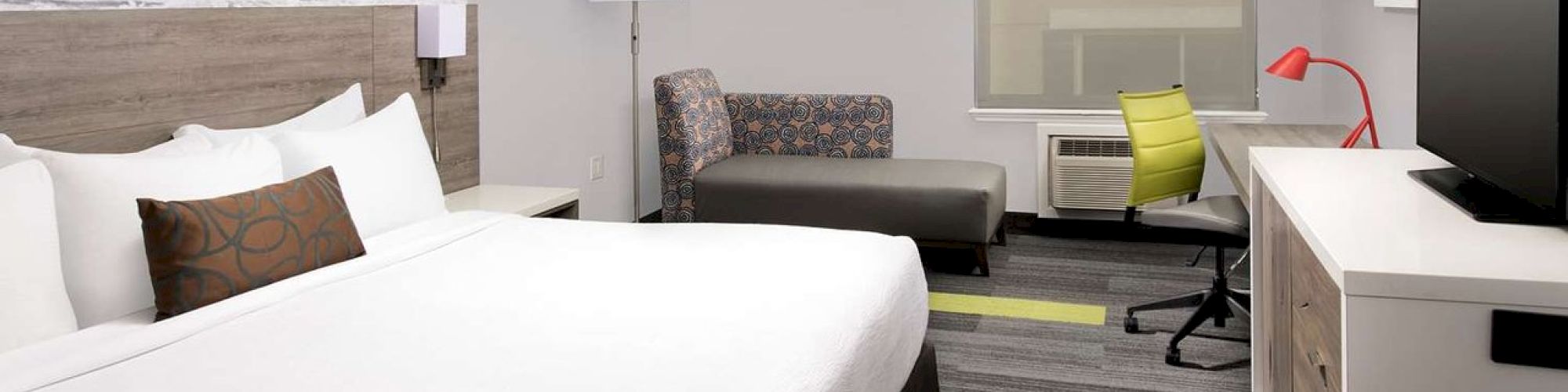 The image shows a modern hotel room with a bed, sofa, desk, TV, and wall art of a surfer. The decor is stylish and contemporary.
