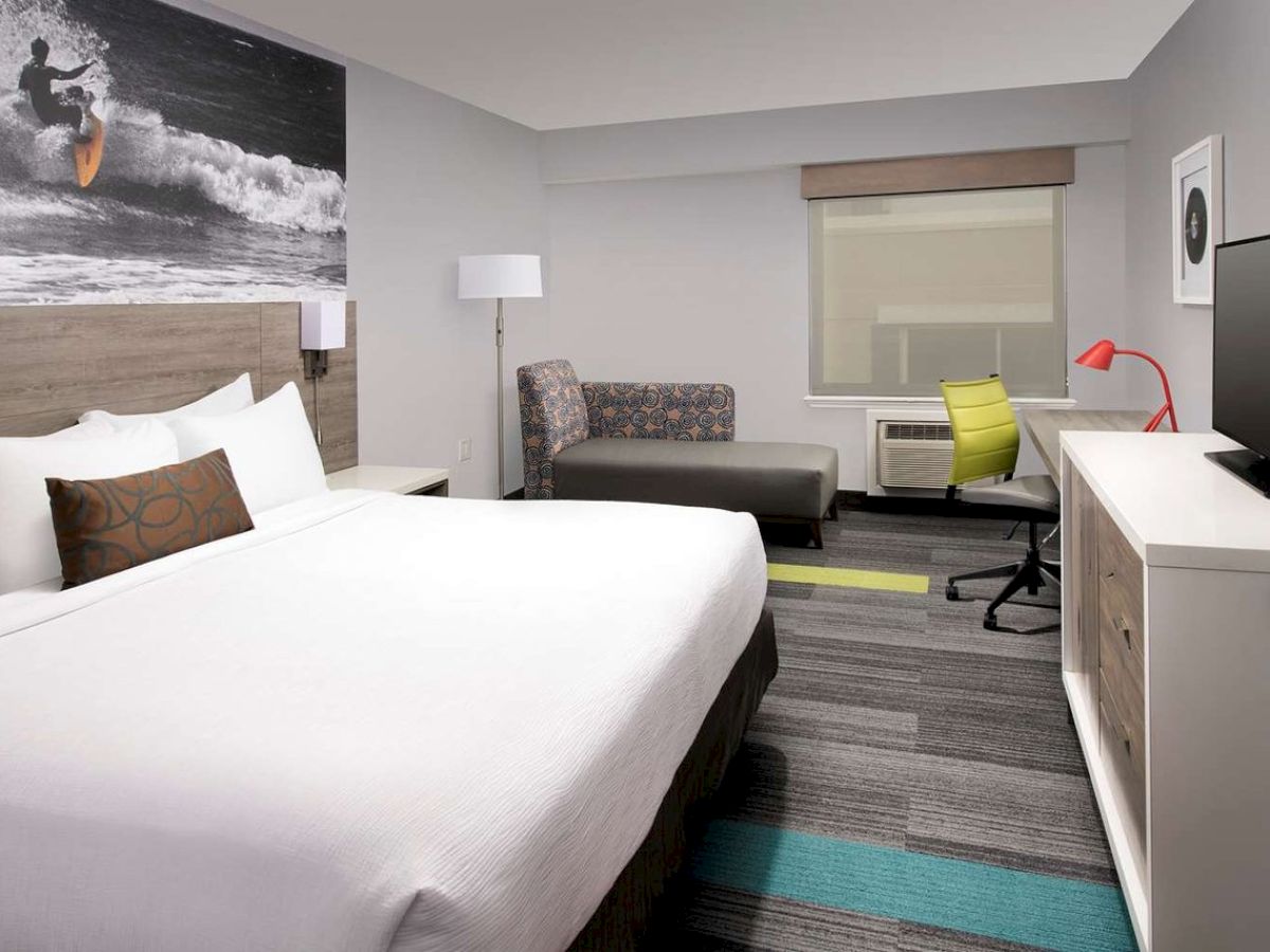 The image shows a modern hotel room with a large bed, TV, desk, and surf-themed artwork above the bed, decorated in neutral and bright tones.