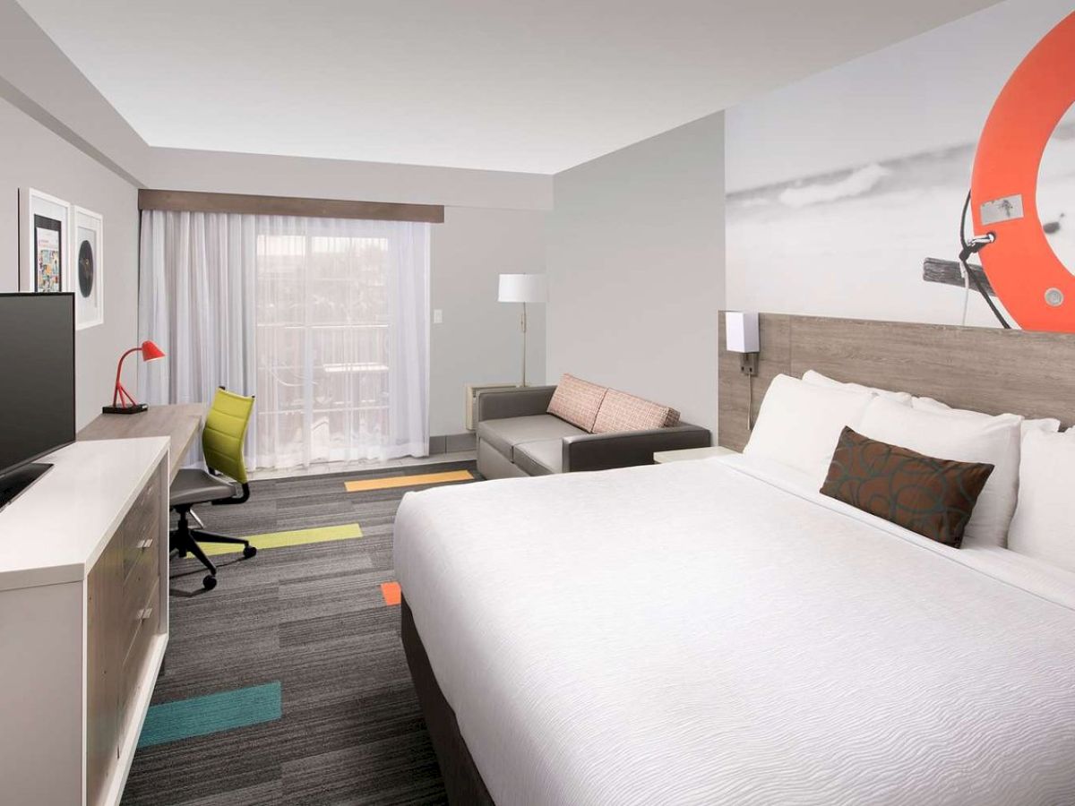 A modern hotel room with a large bed, desk, TV, and couch, featuring a beach-themed wall art and colorful accents throughout.
