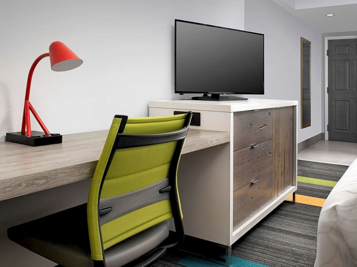 A hotel room with a desk, red lamp, green chair, TV on a dresser, and a bed in a modern setting.