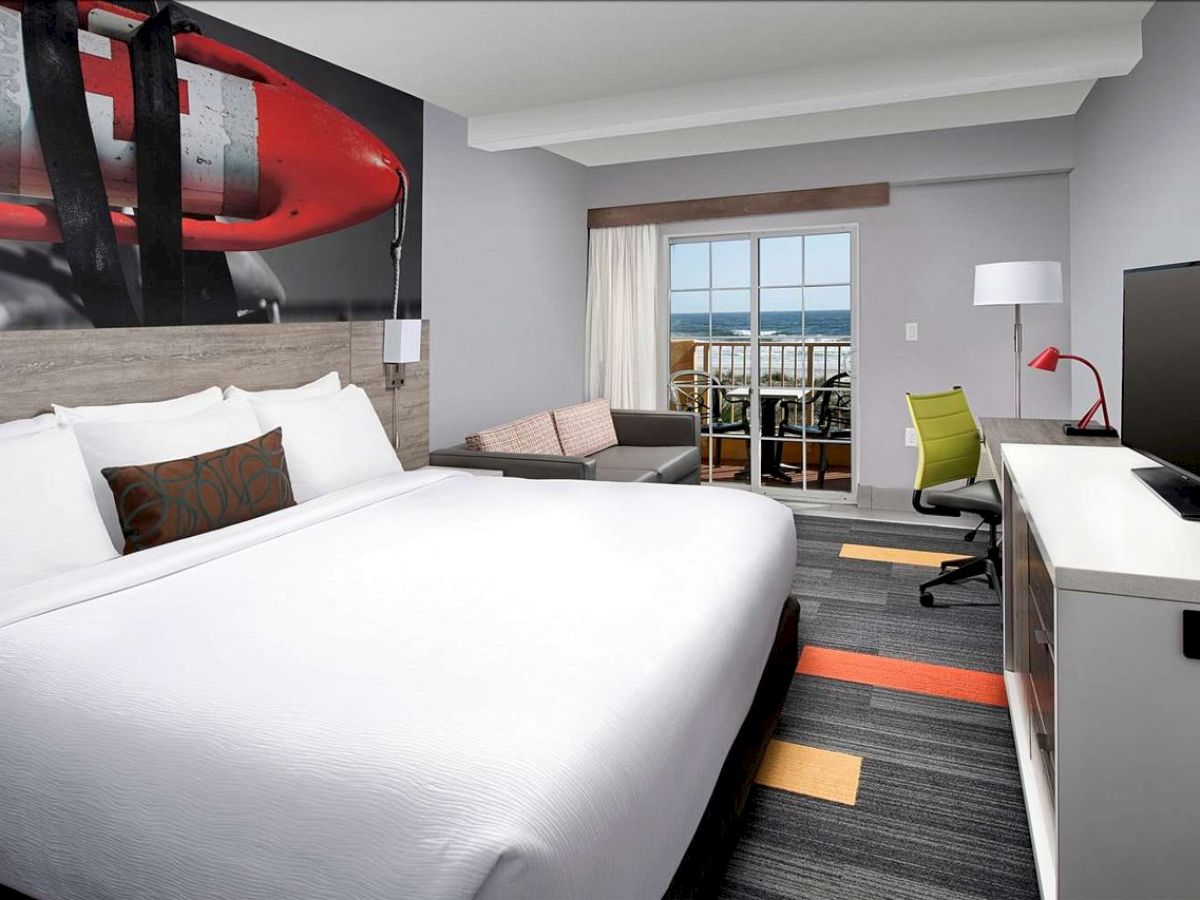 A modern hotel room with a large bed, TV, desk, sofa, and a window with a view, featuring simple decor and colorful accents.