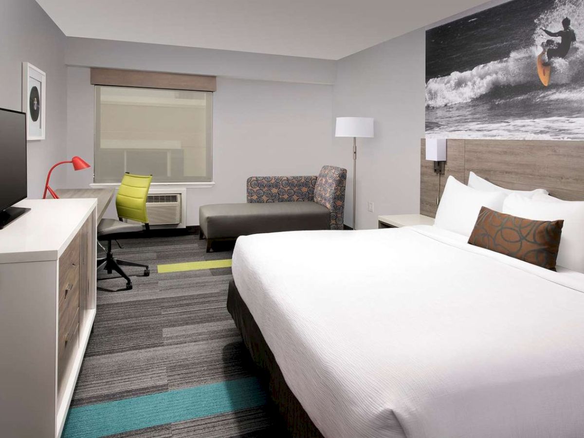 The image shows a modern hotel room with a bed, desk, chair, and a surf-themed artwork on the wall.