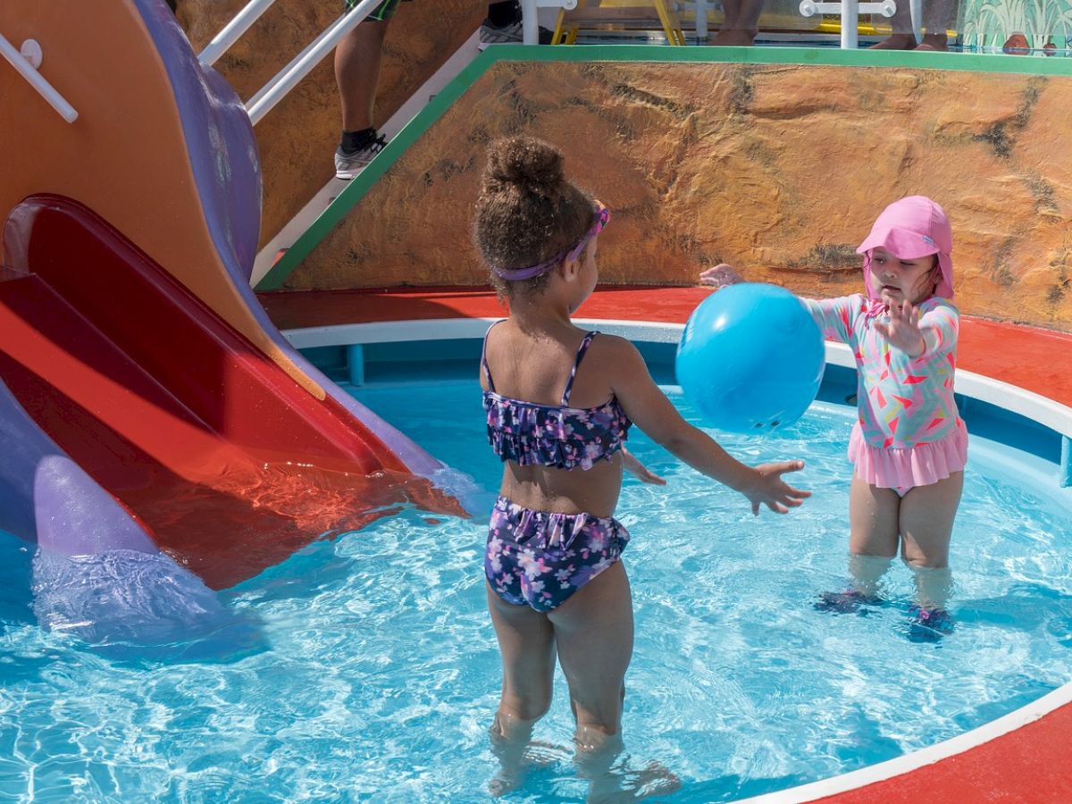 Two children are playing with a ball in a shallow pool next to a small slide, wearing swimsuits and sun hats.
