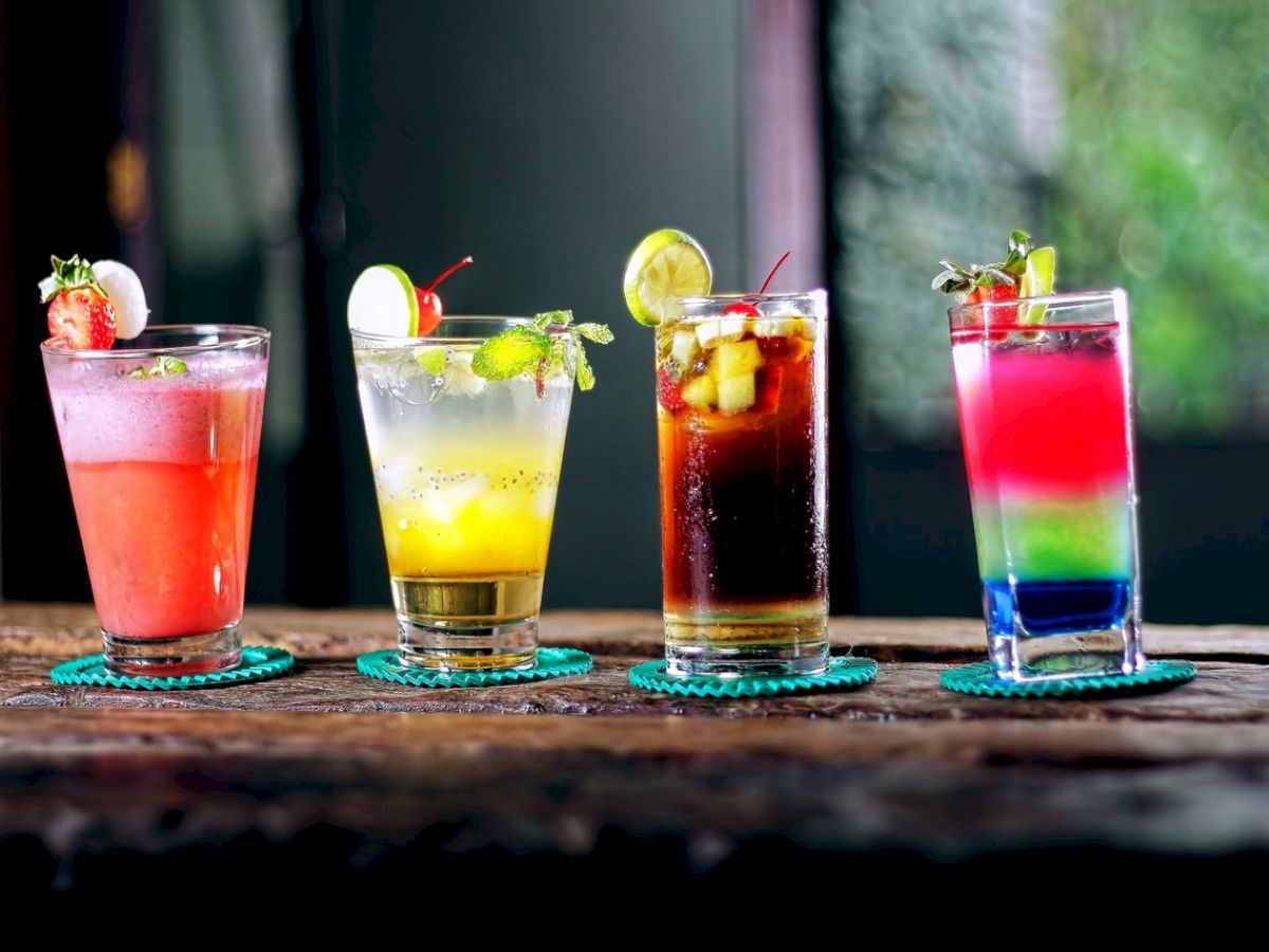 Four colorful cocktails on a wooden table, each garnished with fruit slices and cherries, served in different glasses against a blurred background.