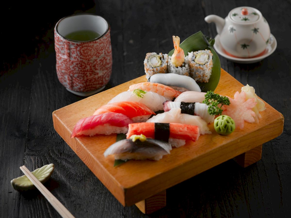 The image shows a sushi platter with assorted sushi, a cup of tea, and a teapot on a dark table, accompanied by chopsticks and a soy sauce dish.