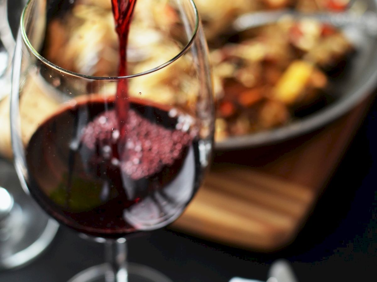 A glass of red wine is being poured from a bottle, with food in the background and napkins on the table.