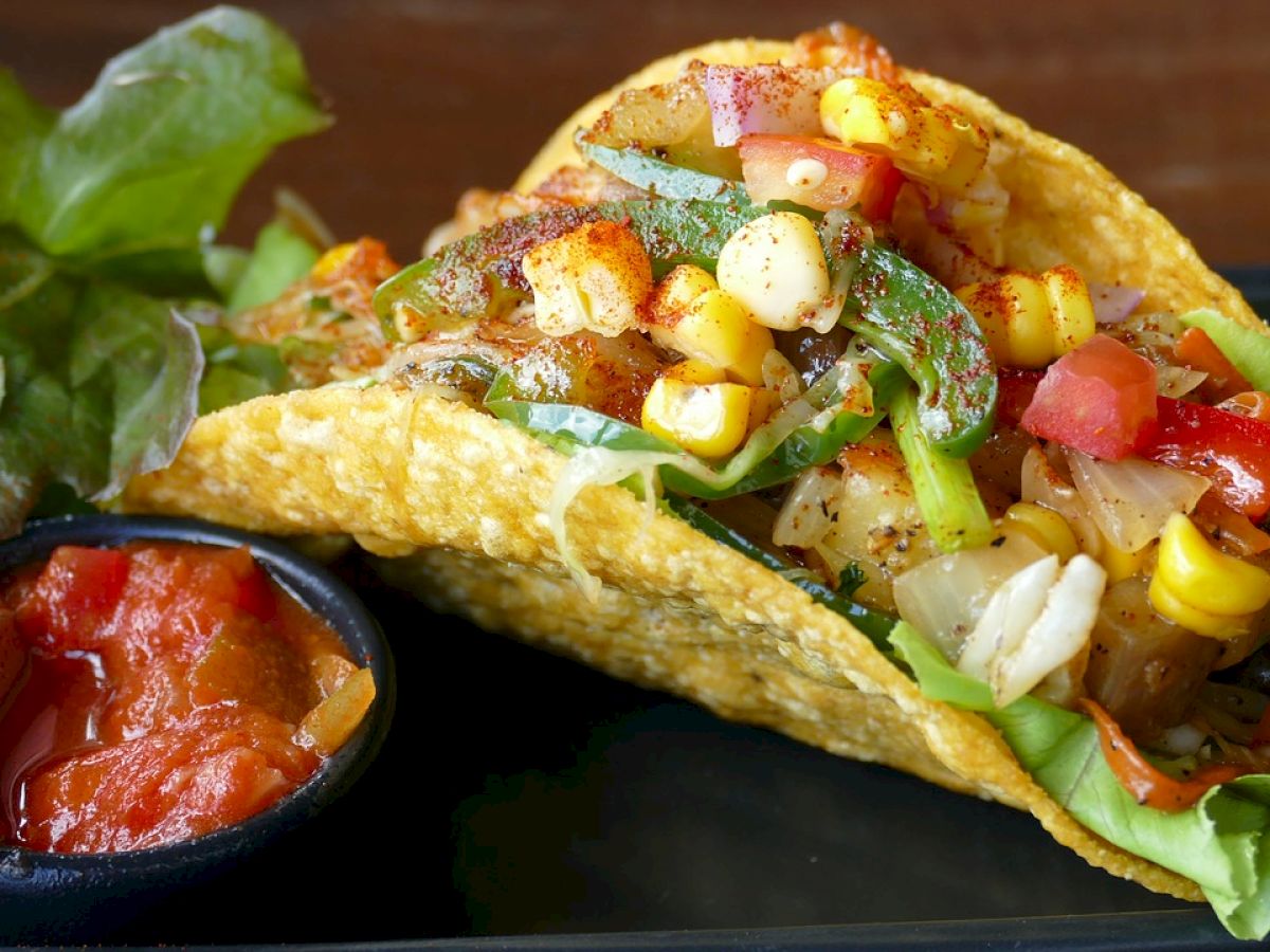 A taco filled with vegetables like corn, lettuce, and tomatoes, accompanied by a small dish of salsa on the side.