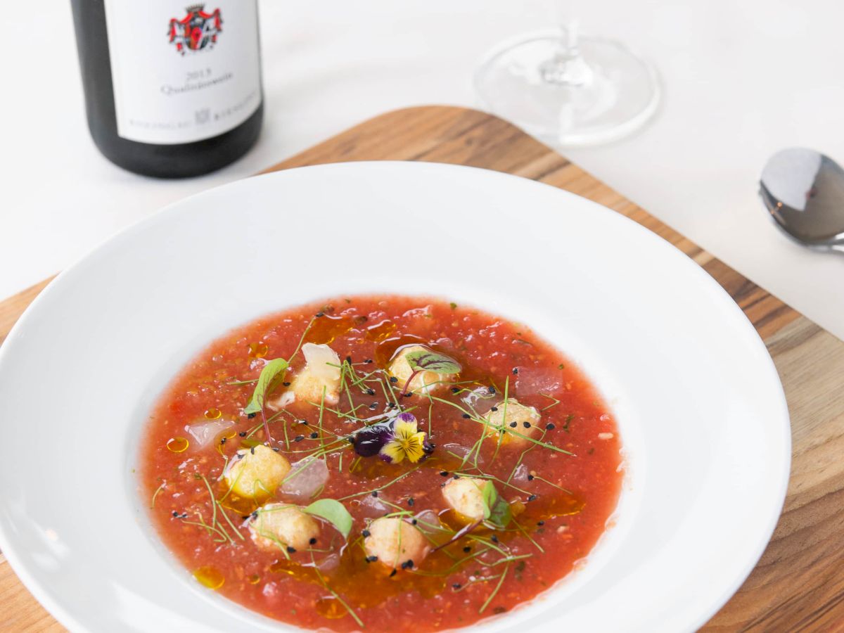 A bowl of tomato soup with garnishes on a wooden board, accompanied by a bottle of wine and a glass.
