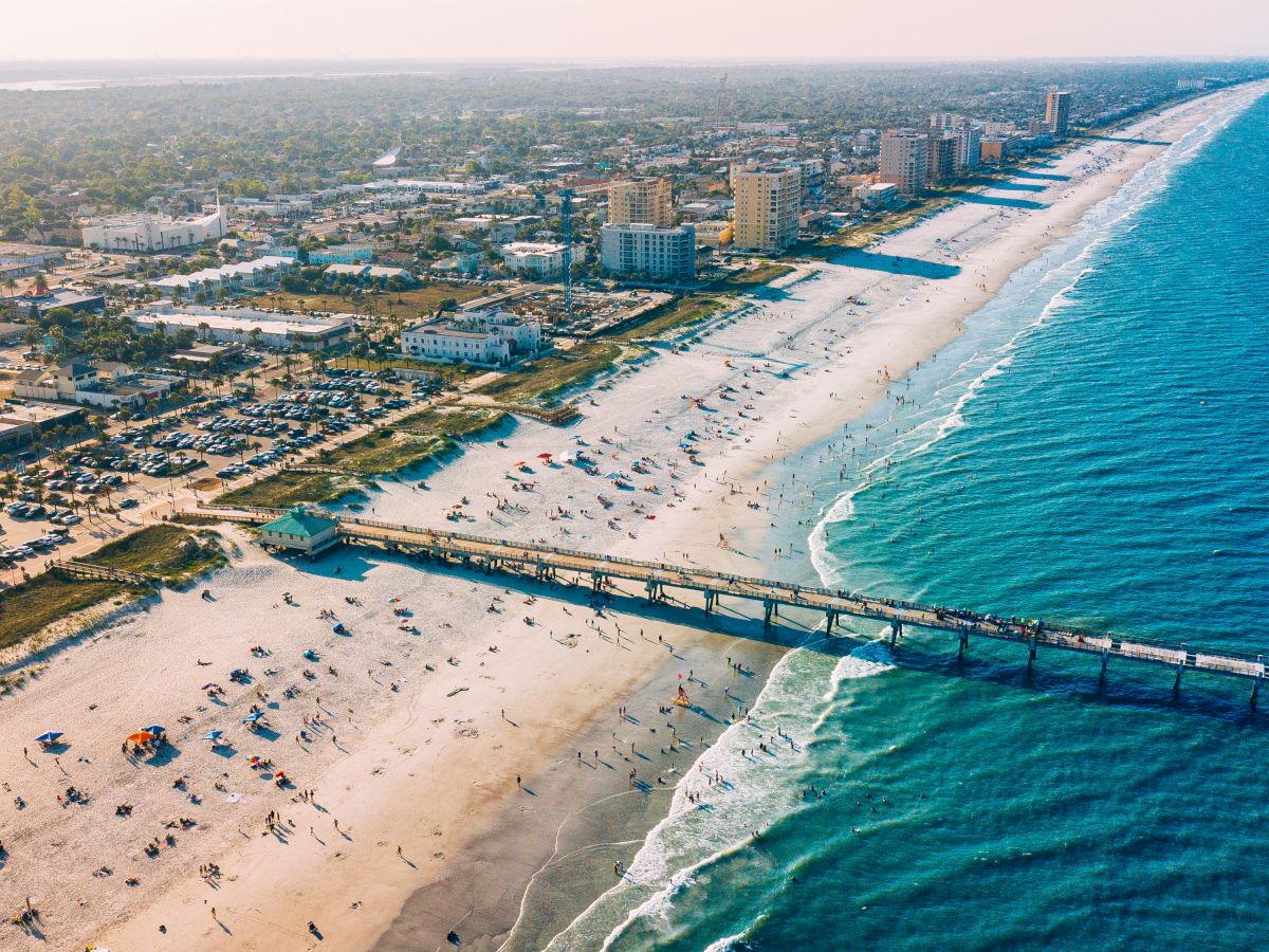 Aerial view of a coastal city with a sandy beach, pier, and ocean, lined with buildings. The beach is dotted with umbrellas and visitors.