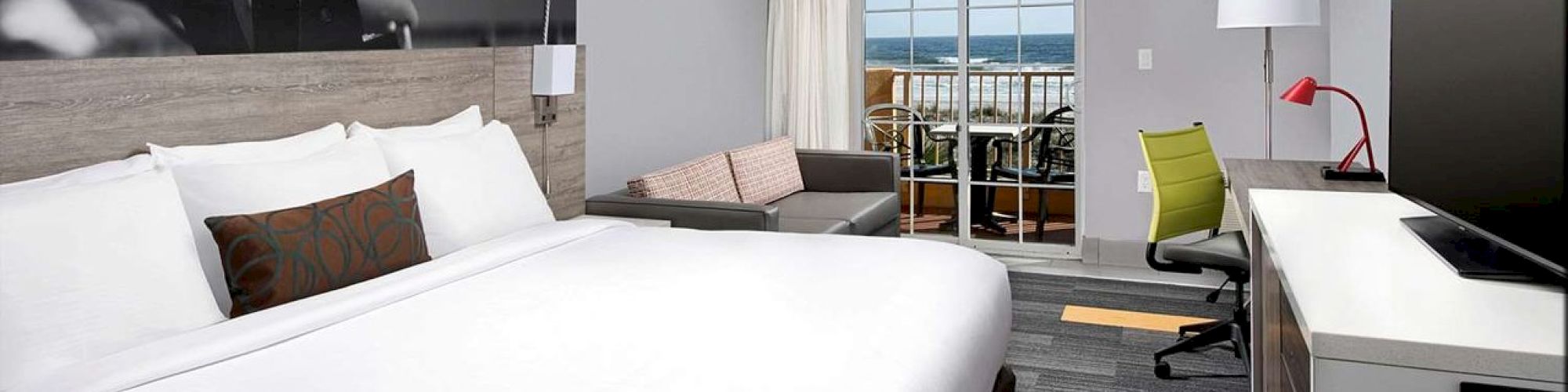 A modern hotel room with a large bed, desk, chair, sofa, balcony with a sea view, and a flat-screen TV on the wall.