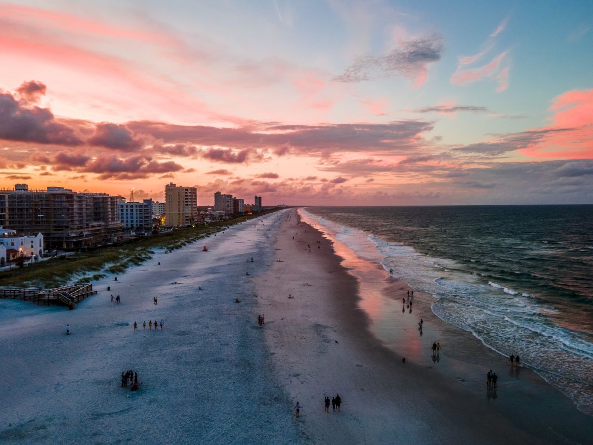 A beach at sunset with pink clouds, buildings on the left, and people walking along the shore, creating a serene coastal scene.