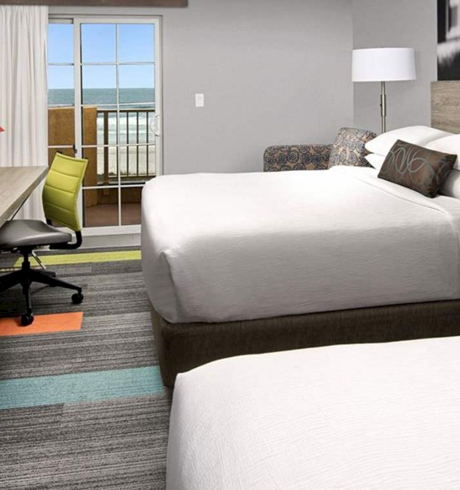 The image shows a hotel room with two large beds, a flat-screen TV, a desk with a chair, and a balcony with an ocean view.
