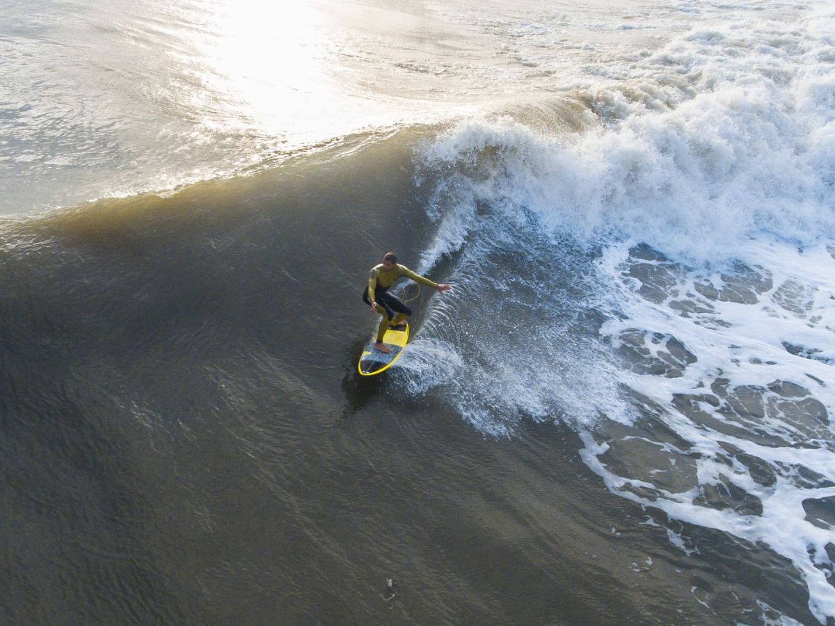 A person surfing on a large wave in the ocean, seen from an aerial perspective; sunlight reflects on the water.