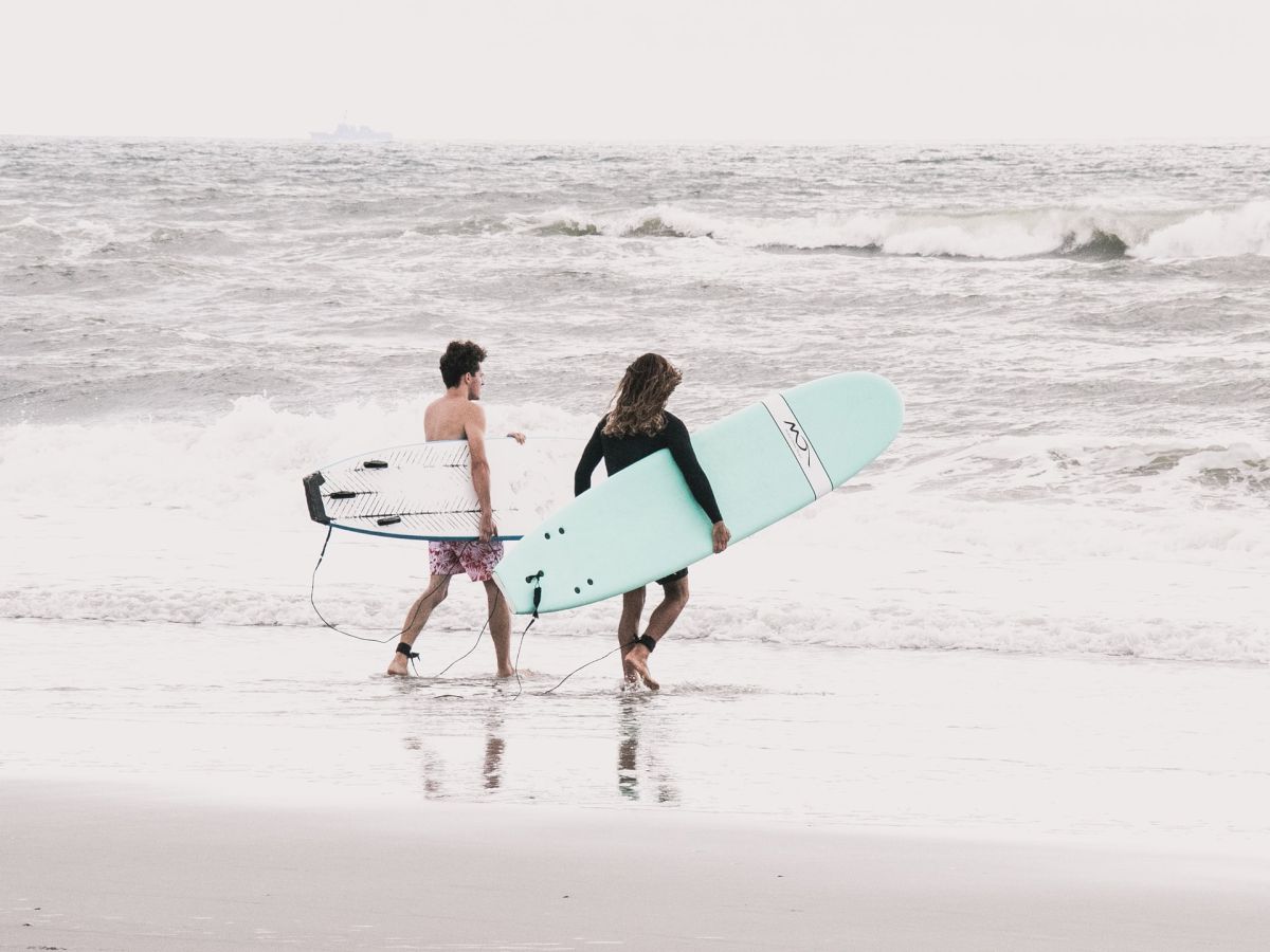 Two people walk along a beach carrying surfboards, with waves in the background.