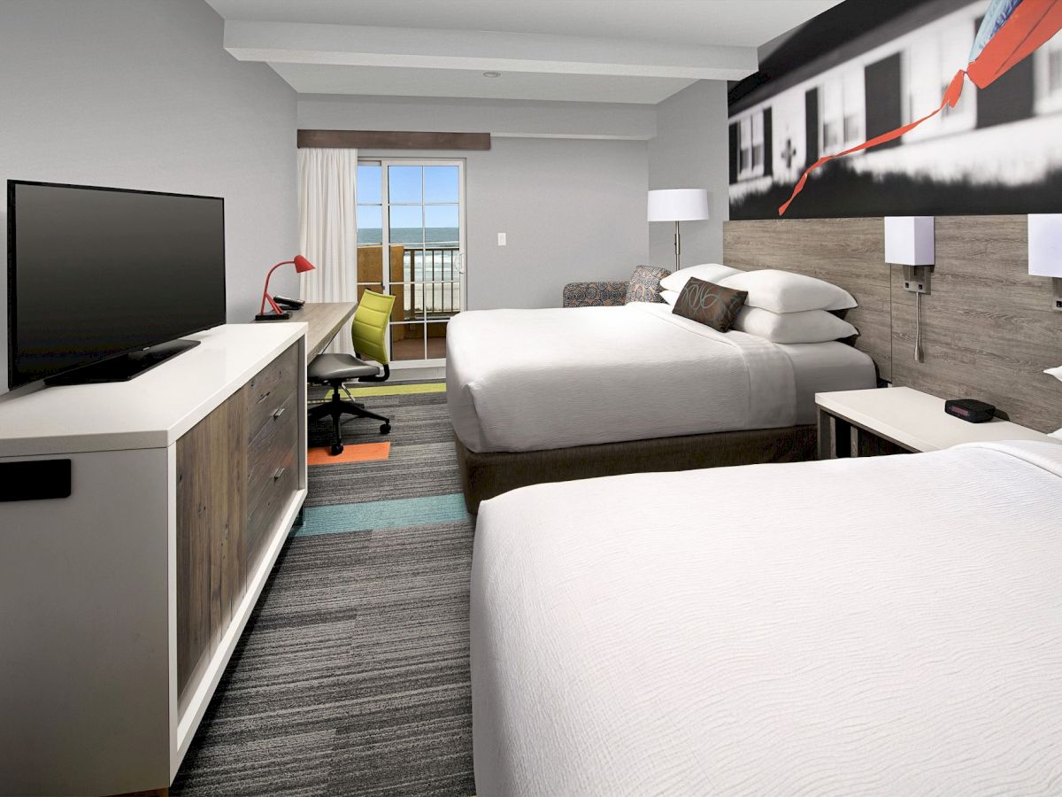 The image shows a hotel room with two beds, a TV on a dresser, a desk with a chair, and a balcony with an ocean view.
