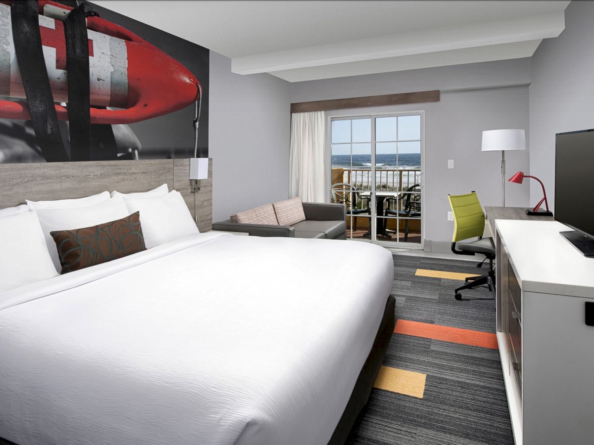 A modern hotel room with a large bed, desk, TV, and seating area, overlooking a balcony with a view of the water.