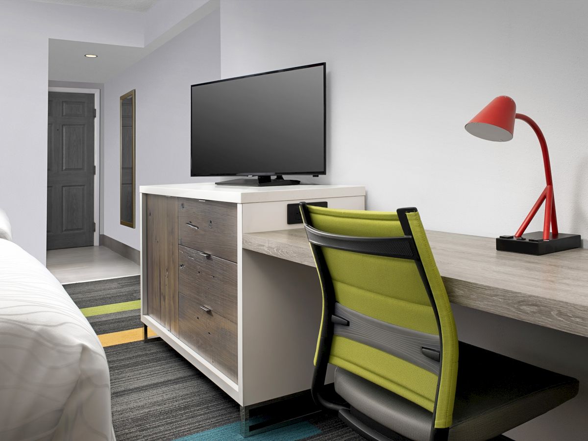 The image shows a hotel room with a bed, TV on a dresser, a desk with a red lamp, and a green office chair on a colorful carpet.