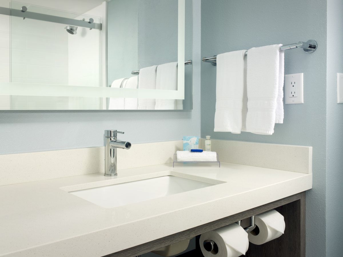 A modern bathroom with a sink, a large mirror, towels on a rack, and toilet paper. The counter is clean and minimalistic in design.