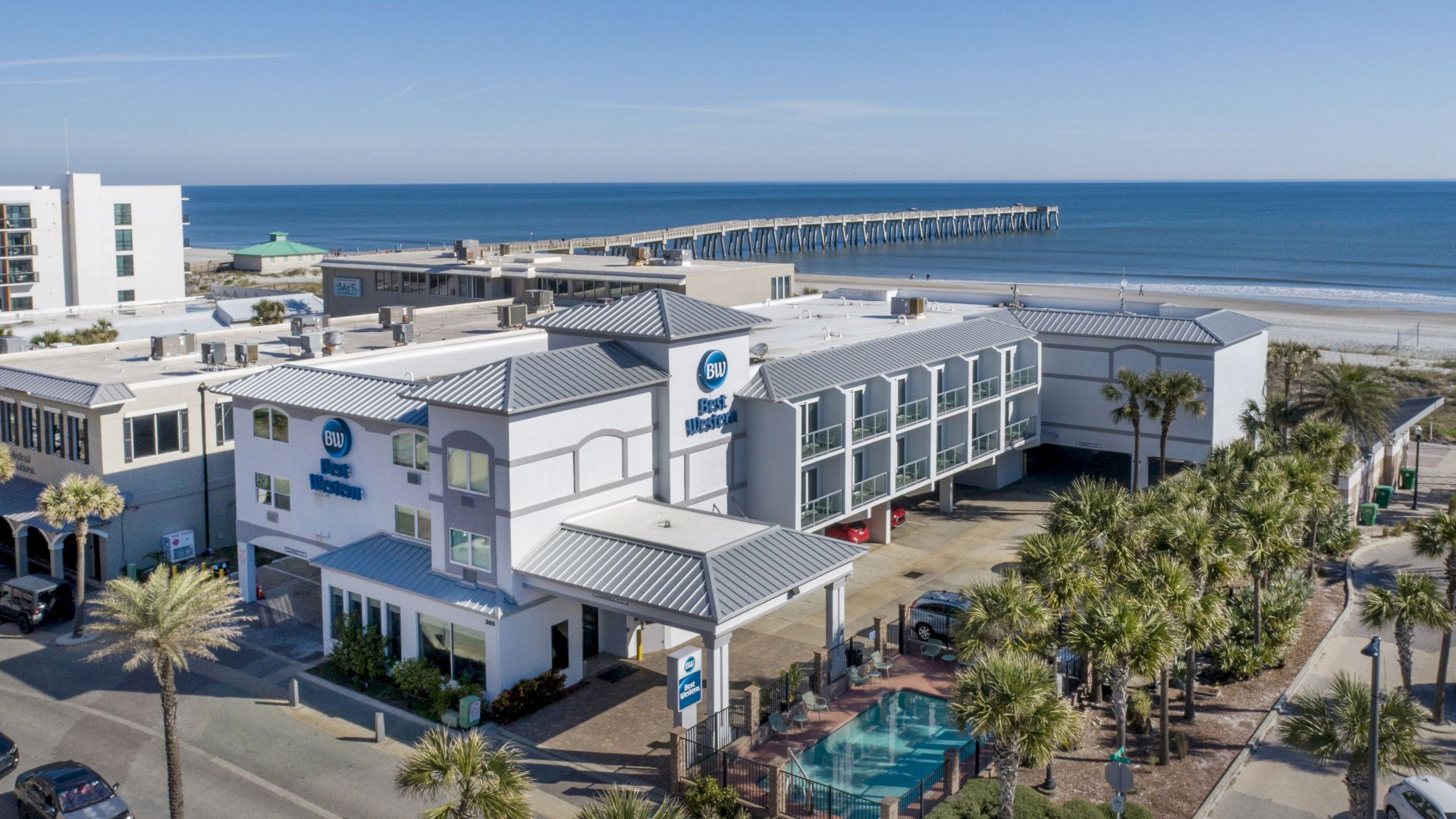A coastal area features hotels, a pool, palm trees, and a long pier extending into the ocean under a clear blue sky.