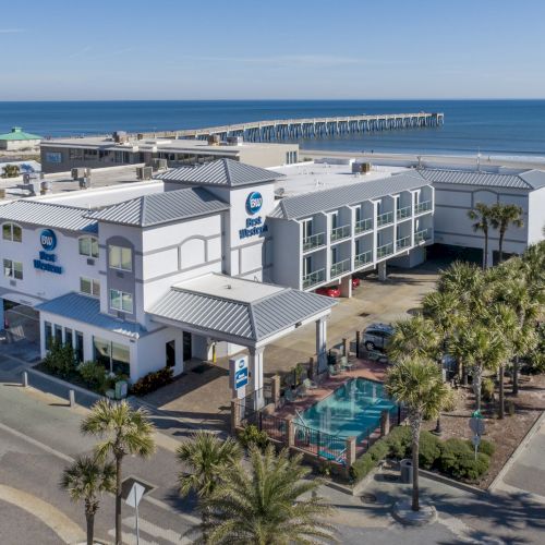 Aerial view of a coastal hotel, surrounded by palm trees and streets, near a pier extending into the ocean under a clear blue sky.