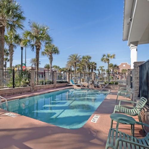 The image shows a swimming pool surrounded by chairs and palm trees, with a clear blue sky in the background.