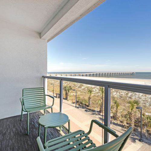 A balcony overlooking a beach with two green chairs and a table, offering a view of the ocean and a pier in the distance.