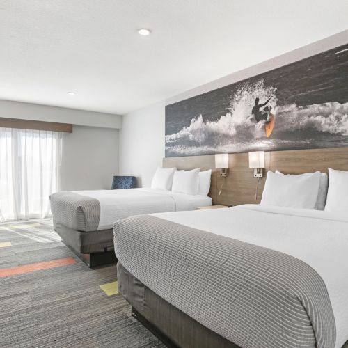 The image shows a hotel room with two beds, a surfing-themed wall art, and modern decor, featuring a TV and natural light from a window.
