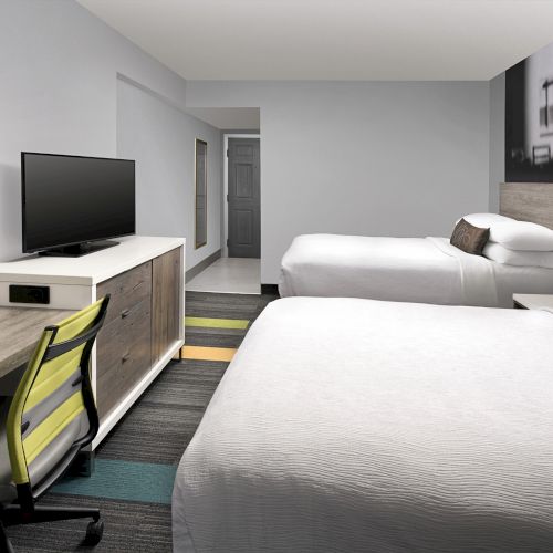A modern hotel room with two beds, a TV, a desk with a red lamp, and a green chair, featuring a colorful floor design.