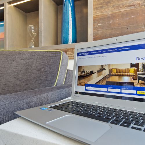 A laptop on a table displaying a hotel booking website next to a gray sofa with green and yellow pillows, in a modern setting.