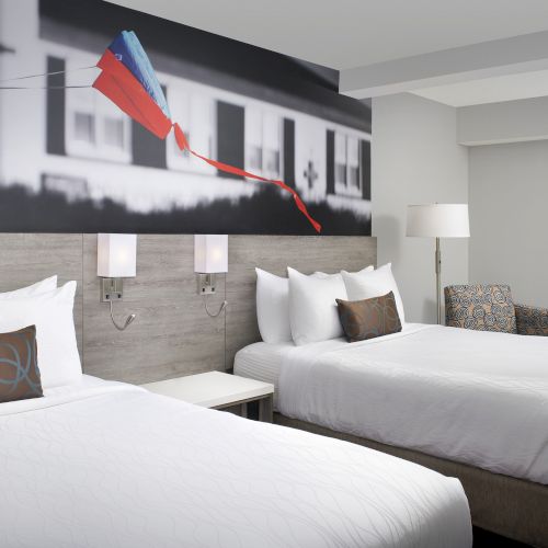 The image shows a hotel room with two double beds, white bedding, and a large wall art piece featuring a colorful kite, creating a cozy space.