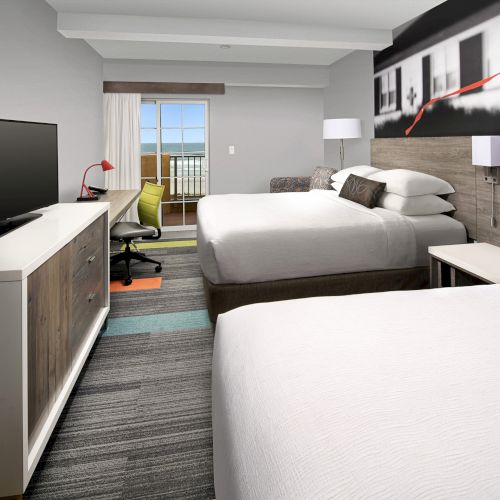 The image shows a modern hotel room with two beds, a TV, a desk, and a balcony view.