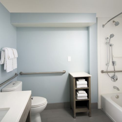 A bathroom with a shower, bathtub, and toilet, featuring gray walls, a towel rack, and a small shelf with stacked towels.