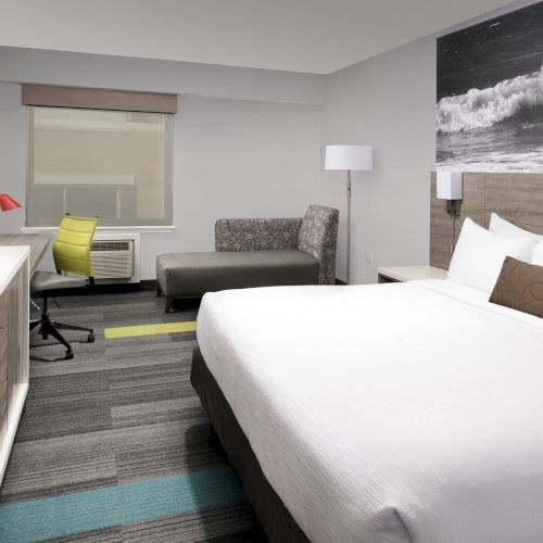 A modern hotel room with a large bed, desk, TV, and artwork of a surfer on the wall. There's a window and seating area.