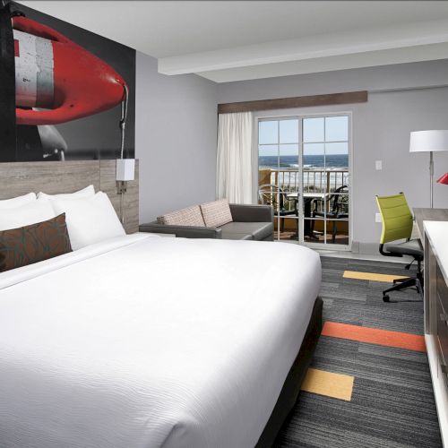 A modern hotel room features a large bed, desk, TV, sofa, and window with a view, decorated in neutral tones with colorful accents.