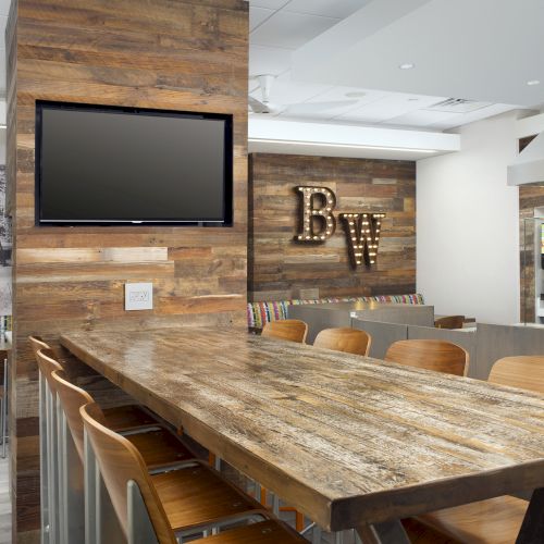 The image shows a modern interior with a wooden table and high chairs, a TV on the wall, and a sign with initials "BW" in the background.